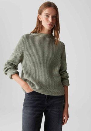 Vrouw in een lichtgroene geribbelde trui met opgerolde mouwen en donkere jeans, staande met een hand in de zak tegen een effen achtergrond.