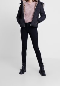 Femme portant une veste matelassée noire avec capuche en fourrure, un pull rose en maille, un pantalon noir skinny et des bottines noires à boucle, debout sur un sol blanc.