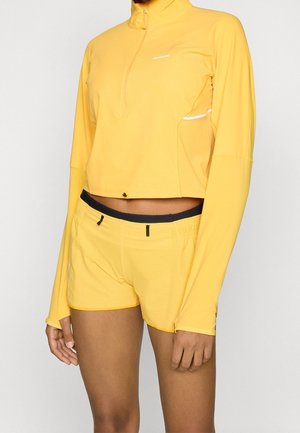 Kobieta ubrana w jaskrawożółtą, długą, krótką bluzkę z ćwiartkowym zamkiem oraz pasujące żółte sportowe szorty na jednolitym tle.