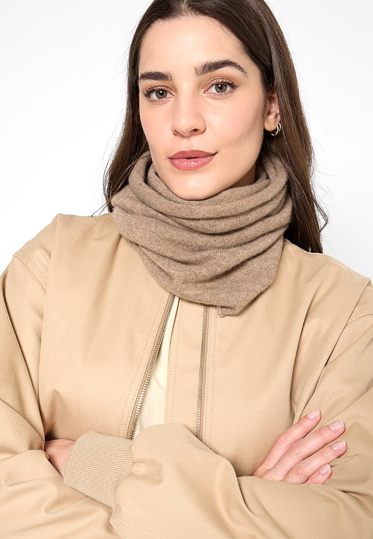 Beżowy pleciony szalik w kształcie nieskończoności owinięty wokół szyi, miękka faktura, w zestawieniu z beżową kurtką bombowcem, z pasującą jasną bluzką pod spodem.