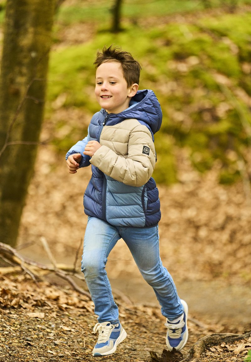 Giacca imbottita con un design a blocchi di colore blu e beige, cappuccio e polsini elastici; abbinata a jeans azzurri chiari e sneakers multicolori.