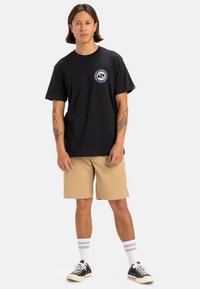 Camiseta de algodón negra con un logo circular en el pecho, combinada con pantalones cortos beige. Los accesorios incluyen zapatillas negras y calcetines blancos con rayas grises.