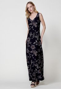 Robe maxi en tissu bleu marine foncé, ornée de motifs floraux et paisley en blanc et rouge. Décolleté en V et sans manches. Sandales à plateforme noires.