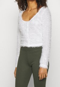 Vit fluffig cardigan med rundad halsringning och långa ärmar, med knappstängning och kort design, kombinerad med gröna leggings.