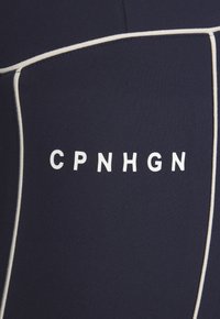 Jūras spēku neoprēna kostīmu materiāls ar baltām akcentiem un tekstu "CPNHGN" treknrakstā. Gluda tekstūra, pieguļošs dizains ar kontrastējošām šuvēm.