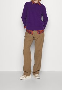 Lila kabelstickad tröja över en mönstrad orange skjorta, kombinerad med beige byxor och krämfärgade sneakers, uppvisad mot en neutral bakgrund.