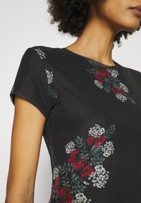 Svart kortärmad topp med blommig broderi i vitt, rött och grönt. Rund halsringning och slät tygtextur, åtsittande design.