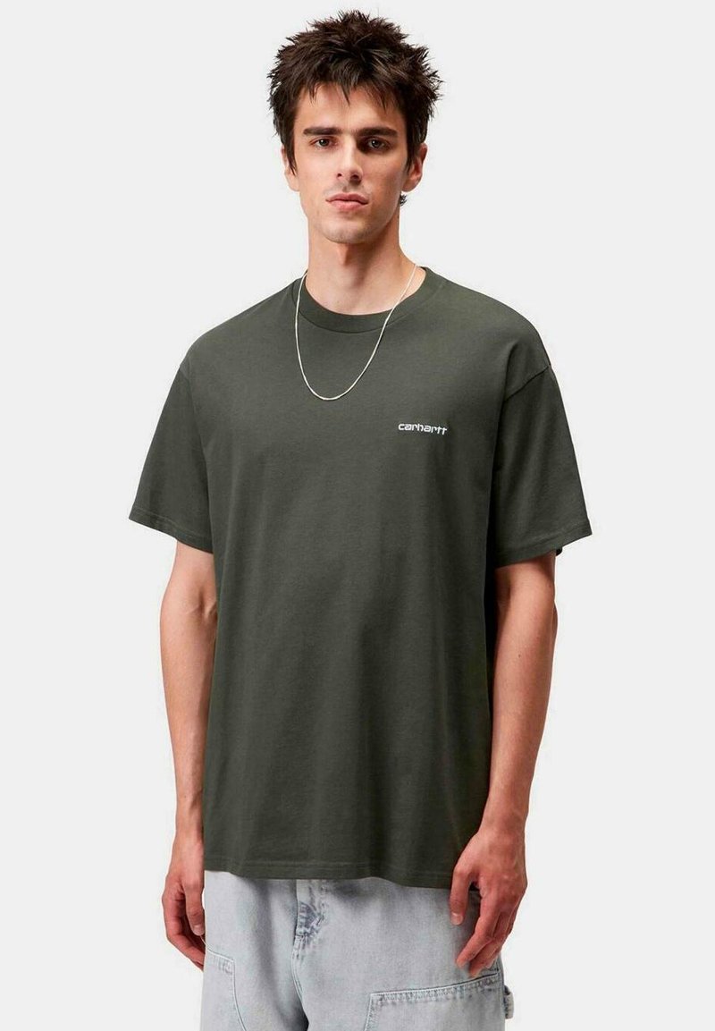 Joven con cabello corto y oscuro que lleva una camiseta Carhartt verde oscuro y pantalones de mezclilla claros contra un fondo liso.