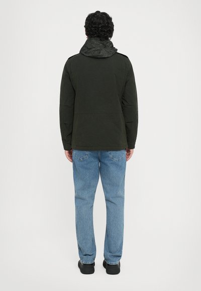 Dunkelgrüne Jacke mit strukturierter Kapuze, Rückansicht. Tragen mit hellblauen Jeans und schwarzen Schuhen, zeigt ein sauberes, einfaches Design.
