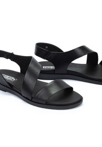 Sandalias negras de cuero con tiras cruzadas, suelas planas, correa ajustable en el tobillo y plantilla texturizada. Logo de la marca visible en la plantilla.