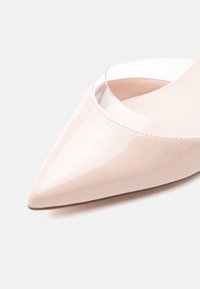 Ballerine en cuir verni nude à bout pointu avec une bordure en vinyle transparent autour de l'ouverture sur un fond clair.