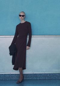 Dlhé, burgundské šaty s dlhými rukávmi, priliehavým telom a splývavou sukňou. Modelka drží tmavý pletený šál. Oblečené s čiernymi slnečnými Okulármi.