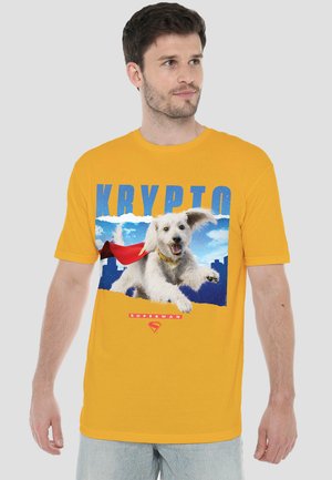 Mann trägt gelbes T-Shirt mit fliegendem weißen Hund mit rotem Umhang, blauem Himmel im Hintergrund und großem blauen Text „KRYPTO“ vorne.