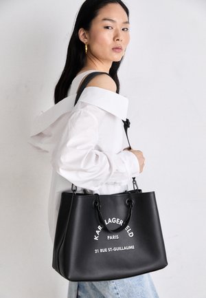 Frau in weißem Off-Shoulder-Top hält eine schwarze Karl Lagerfeld Paris Tragetasche mit weißem Text auf der Vorderseite.