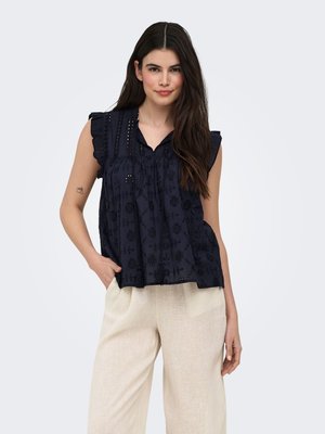 Femme portant un chemisier noir brodé sans manches et un pantalon ample beige clair, debout avec une main dans la poche.