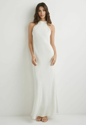 Kobieta stoi przy białej ścianie, ubrana w białą, bezrękawową suknię z wysokim kołnierzem sięgającą podłogi oraz cieliste, odkryte buty na obcasie.