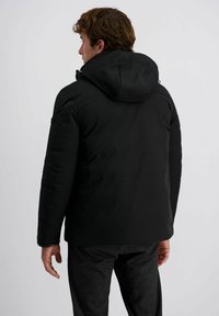 Zwarte geïsoleerde jas met een capuchon, gladde textuur, voorzien van een vooraanstaande rits en elastische manchetten. Draagt bij een geruite broek.