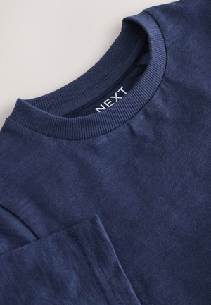 Tumesinise puuvillase T-särgi ümara kaelusega, millel on tekstureeritud kangas ja vasakul pool tasku detail. Kaelusel on brändi silt.