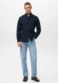 Man staat in een donkerblauwe button-up shirt, lichtblauwe jeans en zwarte leren schoenen tegen een effen achtergrond.