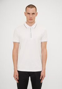 Hvit polo skjorte med svart kant og treknapps lukking. Har "ARMANI EXCHANGE" trykt vertikalt foran. Korte ermer.