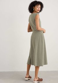 Gestreiftes, ärmelloses Kleid in Grün und Weiß mit einem ausgestellten Midi-Längen-Saum, kombiniert mit braunen Clogs, die ohne Schnürung getragen werden, auf einem neutralen Hintergrund.