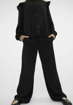 Czarna bluzka z guzikiem z przodu, falbaniaste rękawy i subtelna tekstura, połączona z luźnymi czarnymi spodniami i płaskimi butami.