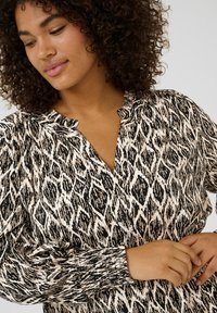 Blouse à motifs noirs et beiges avec un col froncé, des manches longues et des poignets élastiques, présentant un design abstrait texturé.