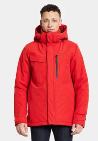 Röd isolerad jacka med hög krage, justerbar huva, flera fickor, blixtlåsstängning och svarta dragkedjeaccenter. Slät textur.