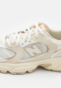 Hvit mesh løpesko med beige detaljer, teksturerte paneler og et fremtredende 'N' logo på siden. Har standard snøring og polstret såle.