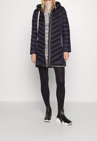 Abrigo acolchado azul marino con capucha, diseño de cuadros en diamante, dos bolsillos laterales, cremallera frontal, usado sobre un vestido gris con estampado y medias negras.