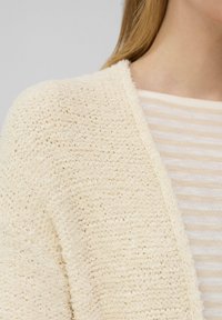 Crème-kleurige gebreide cardigan met een losse structuur. Draagt over een licht gestreept topje met een V-hals en zachte stof.