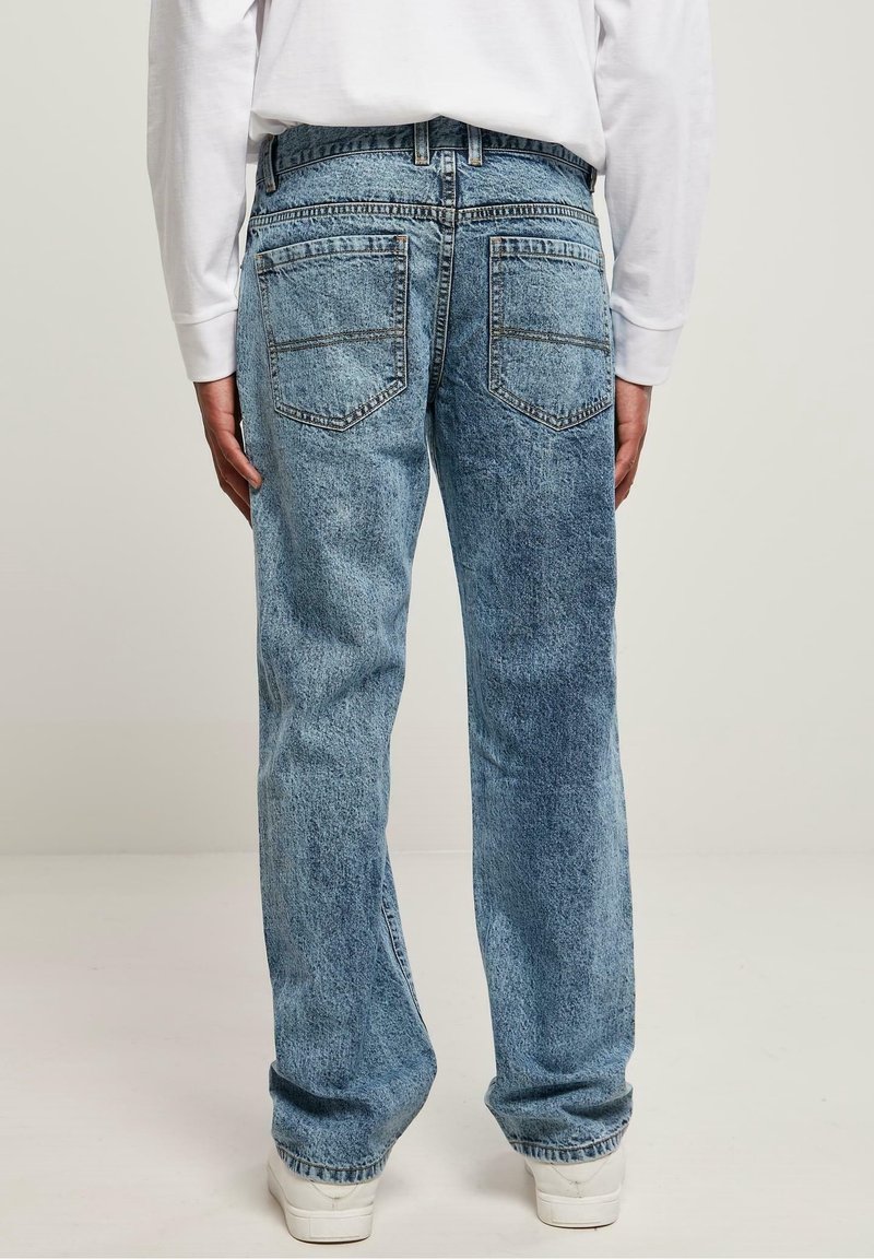 Personne portant un jean bleu droit et des baskets blanches, vue de dos sur un fond uni et clair.