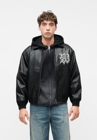 Joven con cabello rizado que lleva una chaqueta de cuero negra sobre una sudadera con capucha negra y jeans azul claro, posando frente a un fondo blanco liso.