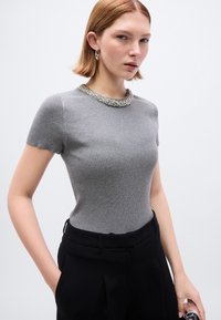 Γκρι κοντομάνικη μπλούζα με ριπ υφή και διακοσμητικό γιακά με χάντρες, εφαρμοστή σχεδίαση και ομαλή υφή. Συνδυάζεται με μαύρο παντελόνι.