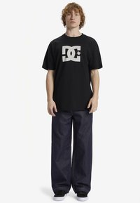Juoda medvilninė marškinėliai su dideliu baltu DC logotipu priekyje, derinama su laisvo silueto tamsaus džinso kelnėmis ir juodais sportiniais bateliais.