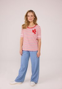 Rot-weiße gestreifte T-Shirt mit einem roten Herzdesign auf der Brust, kombiniert mit hellblauen Schlagjeans und weißen Sneakern.