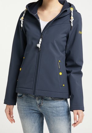 Kobieta ubrana w granatową bluzę z kapturem zapinaną na zamek, z żółtymi guzikami, oraz jasnoniebieskie dżinsy, stojąca z rozluźnionymi ramionami.