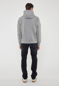 Felpa grigia con cappuccio e vestibilità comoda, con maniche a costine e orlo a costine, abbinata a jeans scuri e sneakers di colore chiaro.