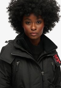 Chaqueta negra aislante con cremalleras prominentes, cuello alto y textura acanalada. Presenta un parche de marca en el hombro y bolsillos laterales.