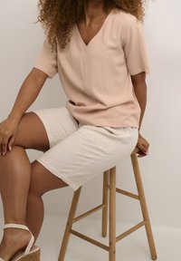 Top rosa cipria a maniche corte con scollatura a V, abbinato a pantaloni pantaloni beige chiaro. Il modello è seduto su uno sgabello di legno, mostrando un tessuto texturizzato.