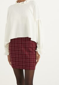 Blusa bianca in tessuto con maniche lunghe e colletto arricciato, abbinata a una gonna a quadri rossa con un motivo a griglia e un design aderente.