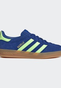 Basket Adidas Gazelle en daim bleu avec trois bandes vert fluo, languette de talon fluo et semelle en gomme translucide.