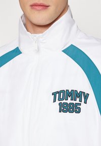 Muž ve bílém zipovém saku s tyrkysovými detaily a vyšitým nápisem "TOMMY 1985" na hrudi, záběr pod nosem.