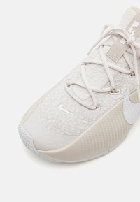 Lys beige atletiksko med tekstureret stofoverdel, flade snørebånd og et hvidt Nike swoosh-logo. Har en polstret, mønstret gummisål.