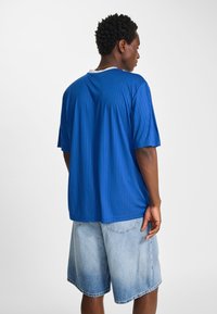 Blå kortärmad skjorta med vertikala ränder, rund halsringning och avslappnad passform, tillsammans med ljusa denimshorts.