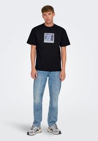 Camiseta negra de manga corta con un estampado gráfico en un cuadrado en la parte delantera. Llevada con jeans de mezclilla azul claro y zapatillas grises.