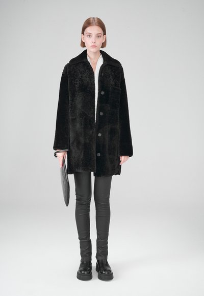 Schwarzer Kunstpelzmantel mit Kragen, Frontknöpfen und einer Brusttasche, kombiniert mit schwarzen Lederleggings und Stiefeln.