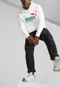 Sudadera blanca con el logo de Puma en rojo y verde, combinada con vaqueros oscuros y zapatillas blancas. Textura suave, capucha ajustable y bolsillo frontal.