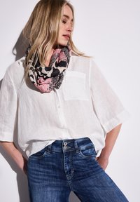 Linnen witte shirt, blauwe denim jeans en een roze en zwart geprint sjaal, gepresenteerd in een casual pose tegen een lichte achtergrond.