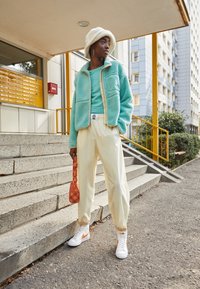 Turkos fleecejacka över en turkos topp, krämfärgade byxor och vita sneakers med orange detaljer. Håller en mönstrad orange väska.